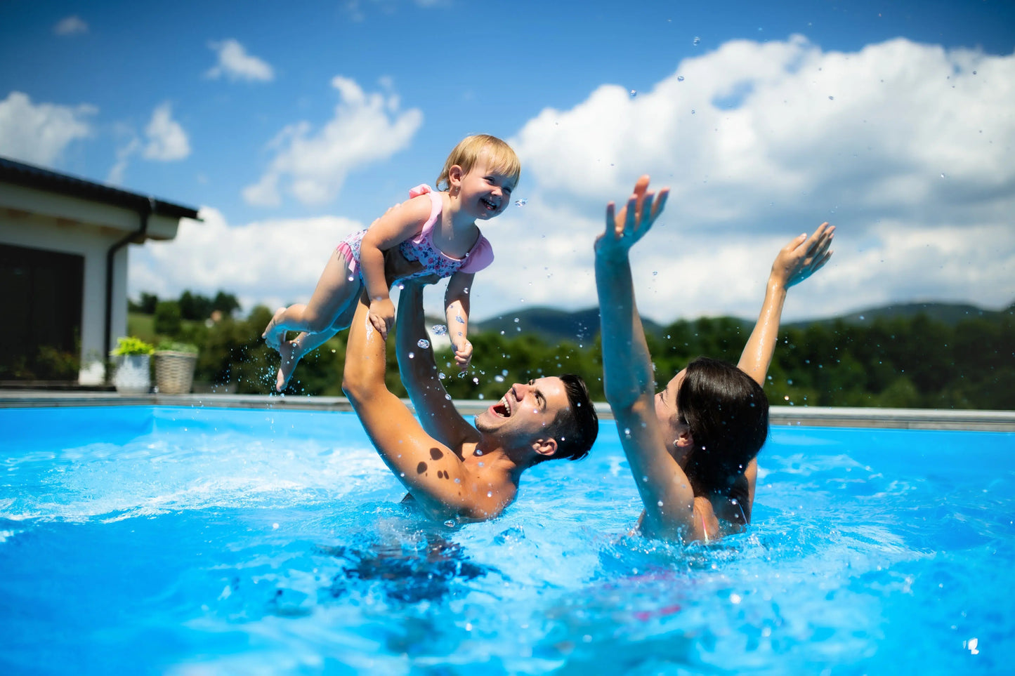 Happy family playing in a pool, featuring a toddler in a pink swimsuit being lifted by a smiling parent.